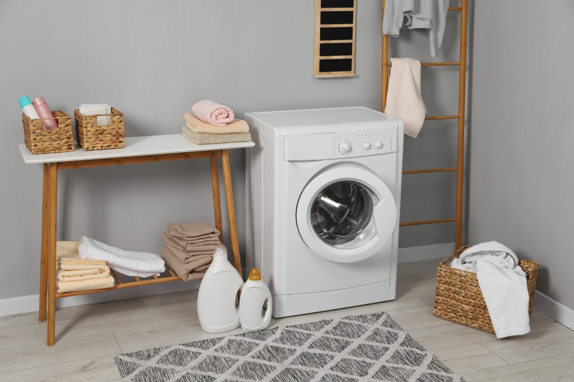 Une petite buanderie avec lave-linge blanc, paniers tissés, serviettes pliées et tapis gris