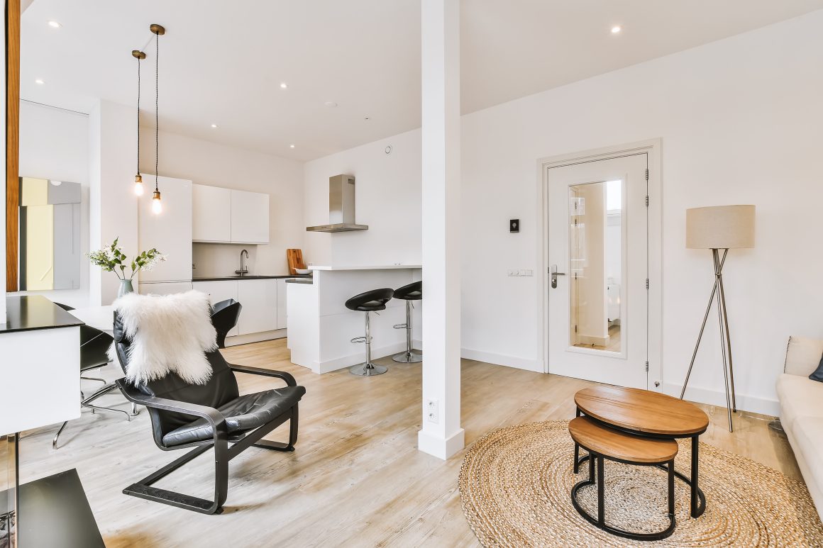 Salon lumineux avec cuisine ouverte, fauteuil noir avec coussin fourrure, tables gigognes sur tapis et lampadaire