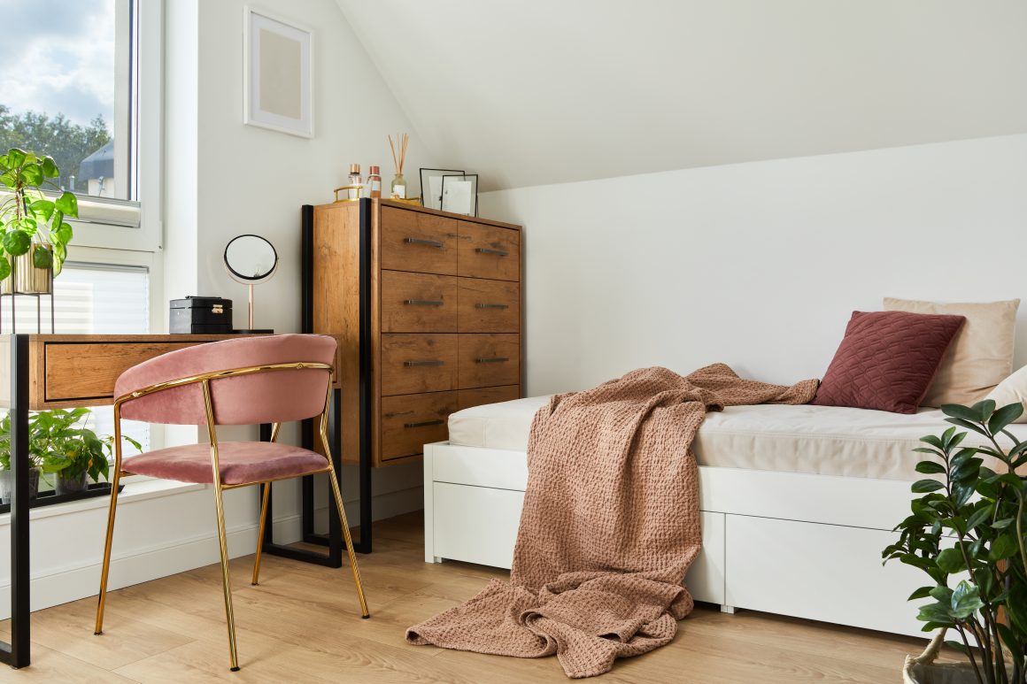 Chambre minimaliste incluant lit blanc, commode en bois, bureau épuré, fauteuil rose en velours et plantes.