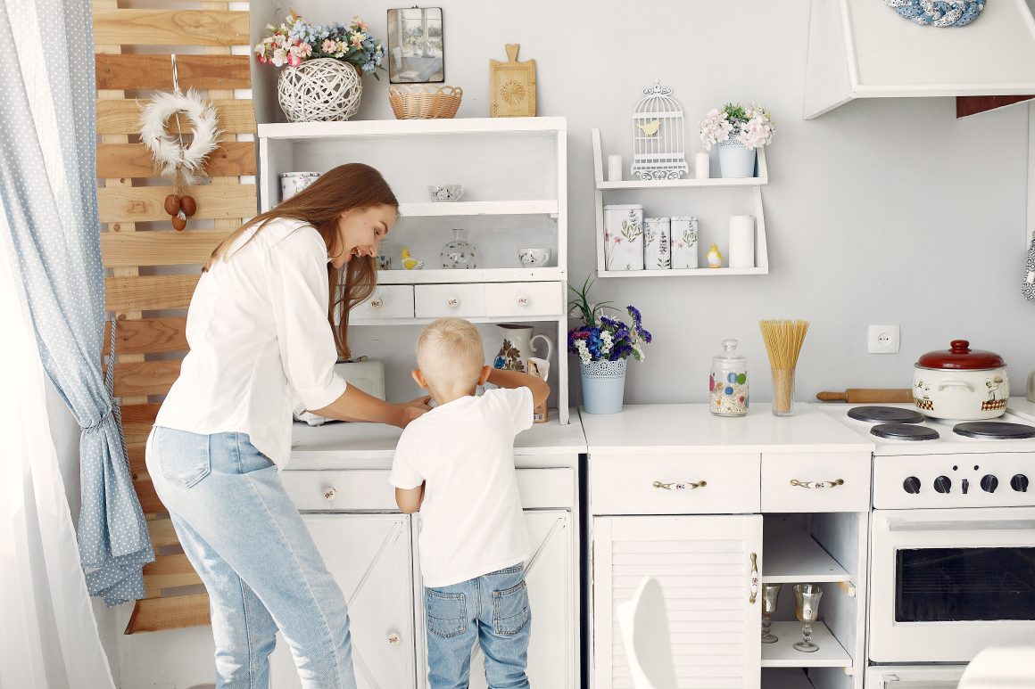 Une mère aide son fils dans une cuisine rustique incluant pots, rangements et étagères ouvertes.