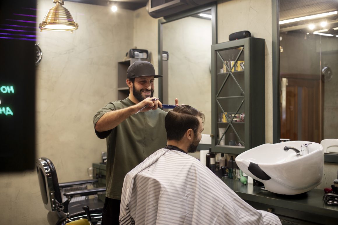 Un coiffeur et un client dans un salon moderne incluant mobilier vert foncé, miroir large et lampe cuivrée