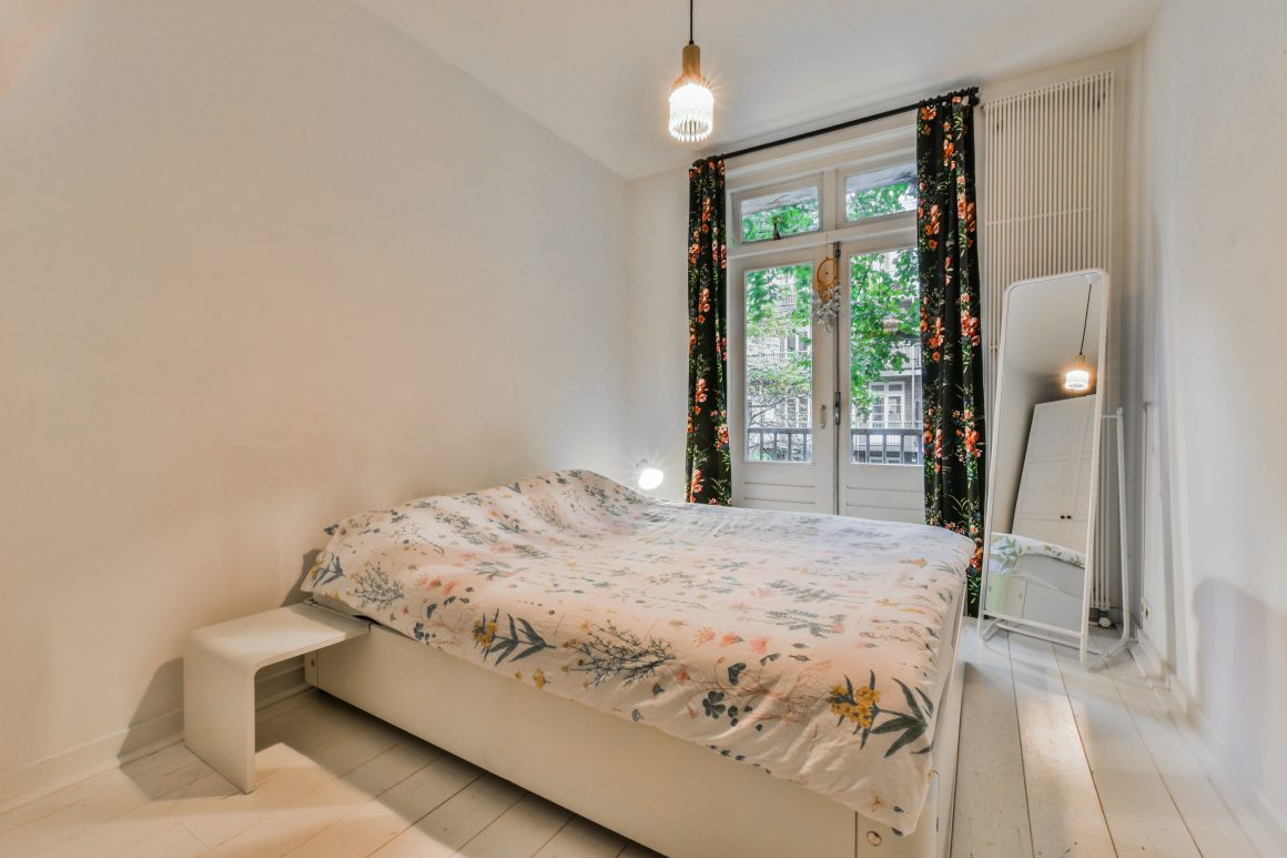 Chambre avec literie florale, grandes portes-fenêtres, rideaux sombres à motifs, miroir et parquet blanc
