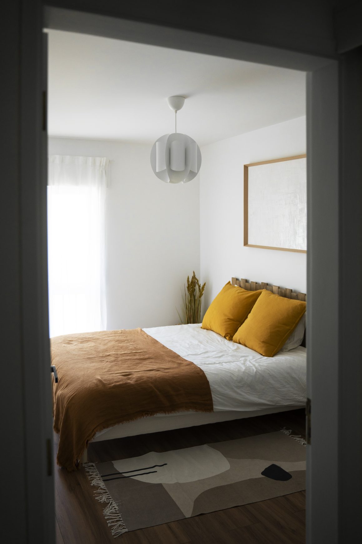 Chambre minimaliste incluant literie cosy, coussins moutarde, tapis abstrait et suspension blanche