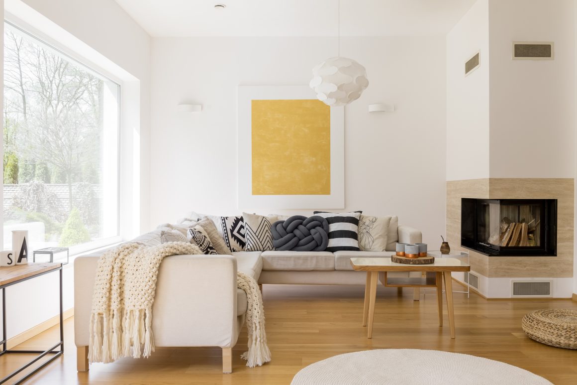 Canapé beige en L, cheminée, table basse bois, tapis rond, murs blancs et lumière naturelle