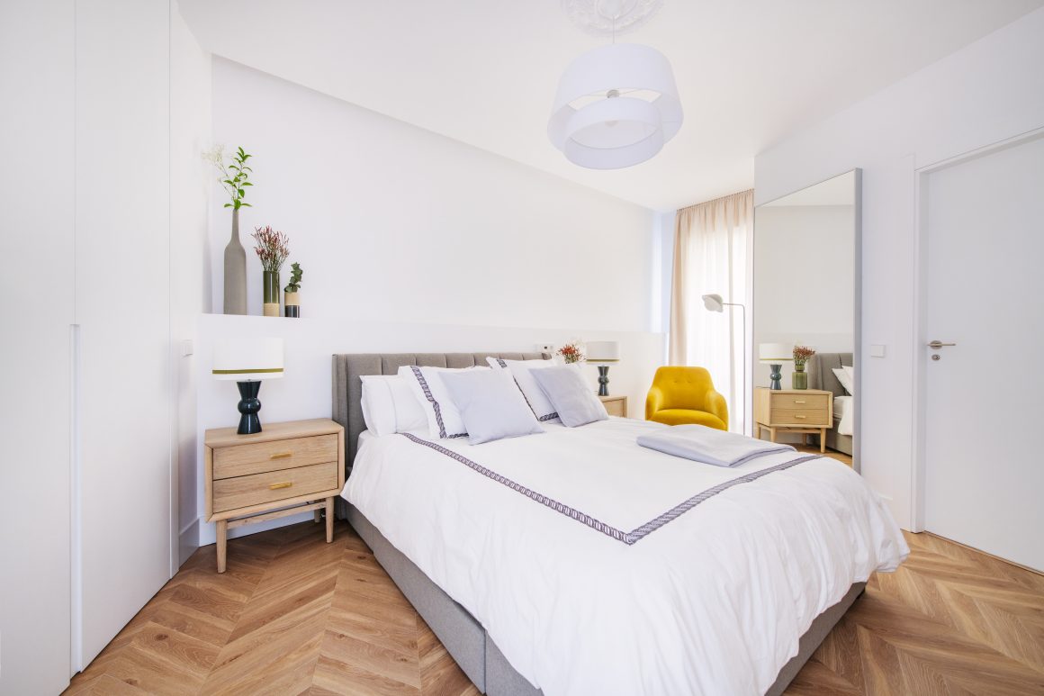 Chambre moderne avec lit double fauteuil moutarde, tables de chevet bois, lampadaires, plantes et miroir