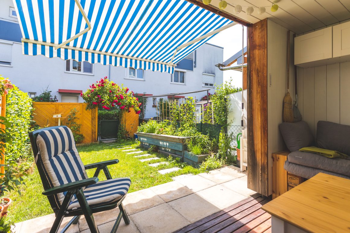 Jardin maison avec terrasse couverte incluant fauteuil rayé, table en bois, une banquette et un store banne
