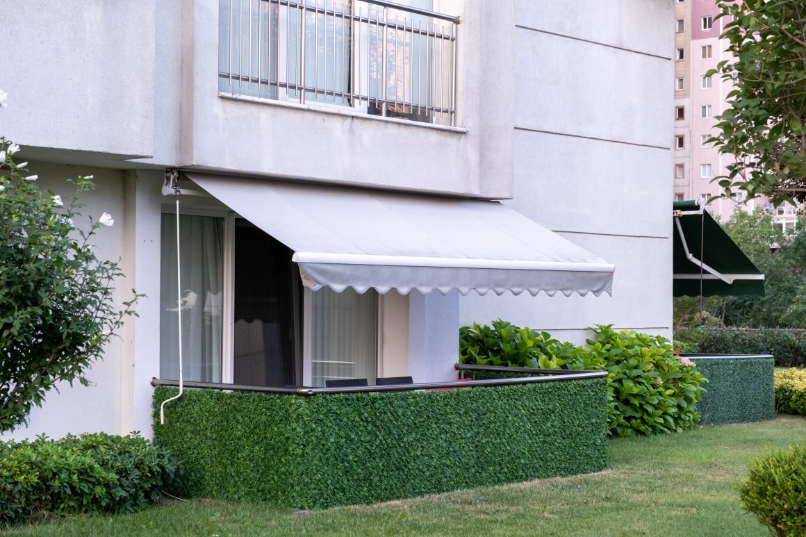 Balcon rez-de-jardin avec store blanc déployé, haie artificielle verte, pelouse soignée et buissons.
