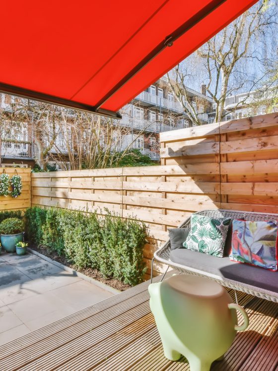 Coin terrasse accueillante : canapé tressé, coussins tropicaux, plantes et auvent rouge
