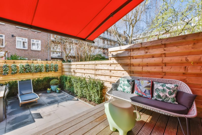 Coin terrasse accueillante : canapé tressé, coussins tropicaux, plantes et auvent rouge