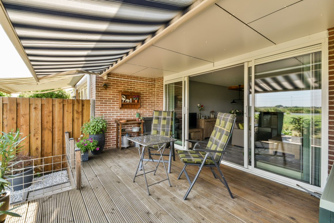 Terrasse en bois avec chaises à carreaux, table, plantes en pots, store rayé et grande baie vitrée