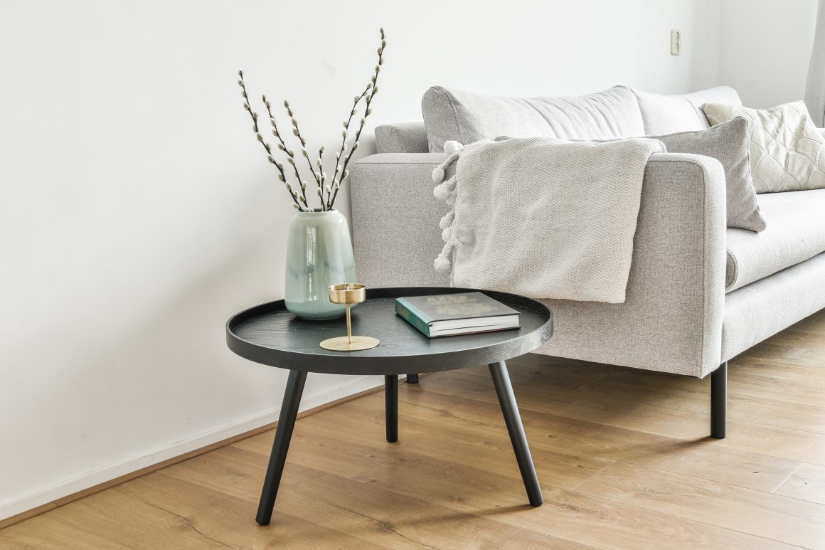 Table basse noire ronde devant un canapé gris, vase vert pâle avec branches, bougeoir doré et livre.