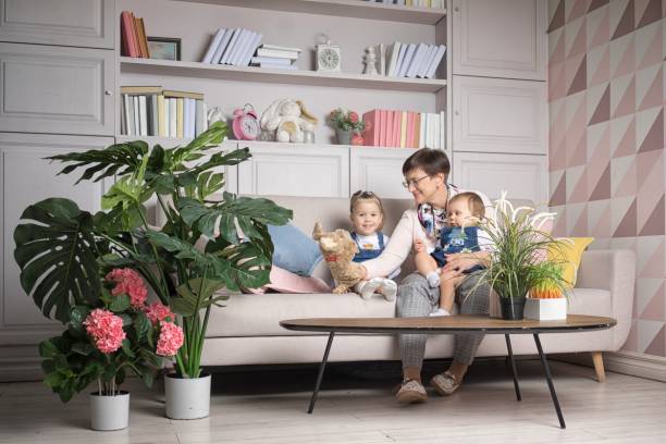Une mère souriante sur un canapé avec deux jeunes enfants, peluches et grandes plantes