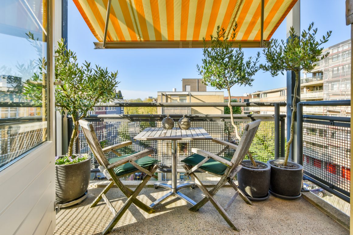 Petit balcon incluant une table en bois, des chaises, des plantes en pots et un store rayé jaune