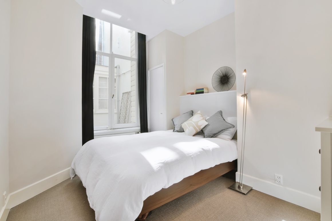 Chambre lumineuse et épurée aux tons neutres avec lit cosy, lampadaire et grande fenêtre.
