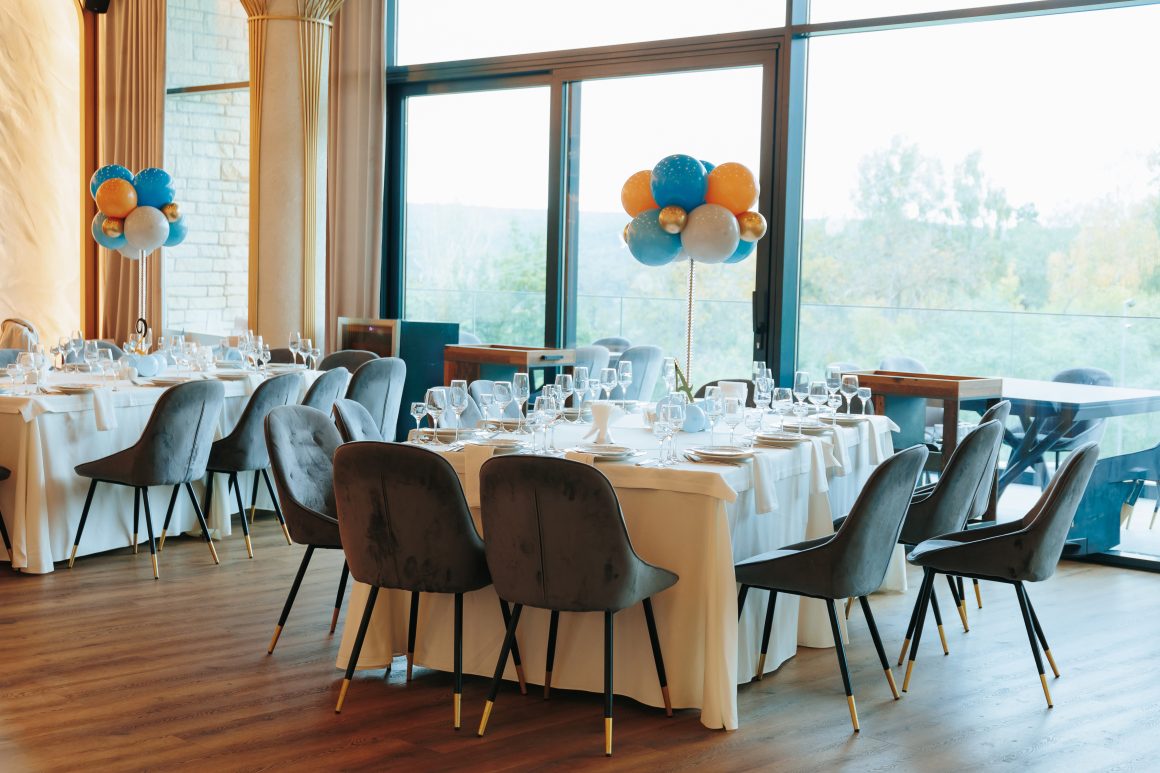 Salle de restaurant décorée avec tables incluant nappes blanches, vaisselle raffinée et bouquets de ballons colorés.