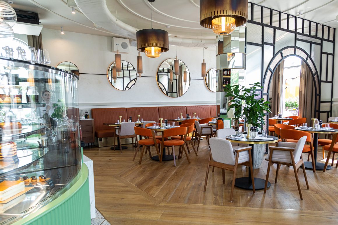 Restaurant aménagé avec chaises orange, banquettes, tables dressées, miroirs ronds, suspensions et plantes