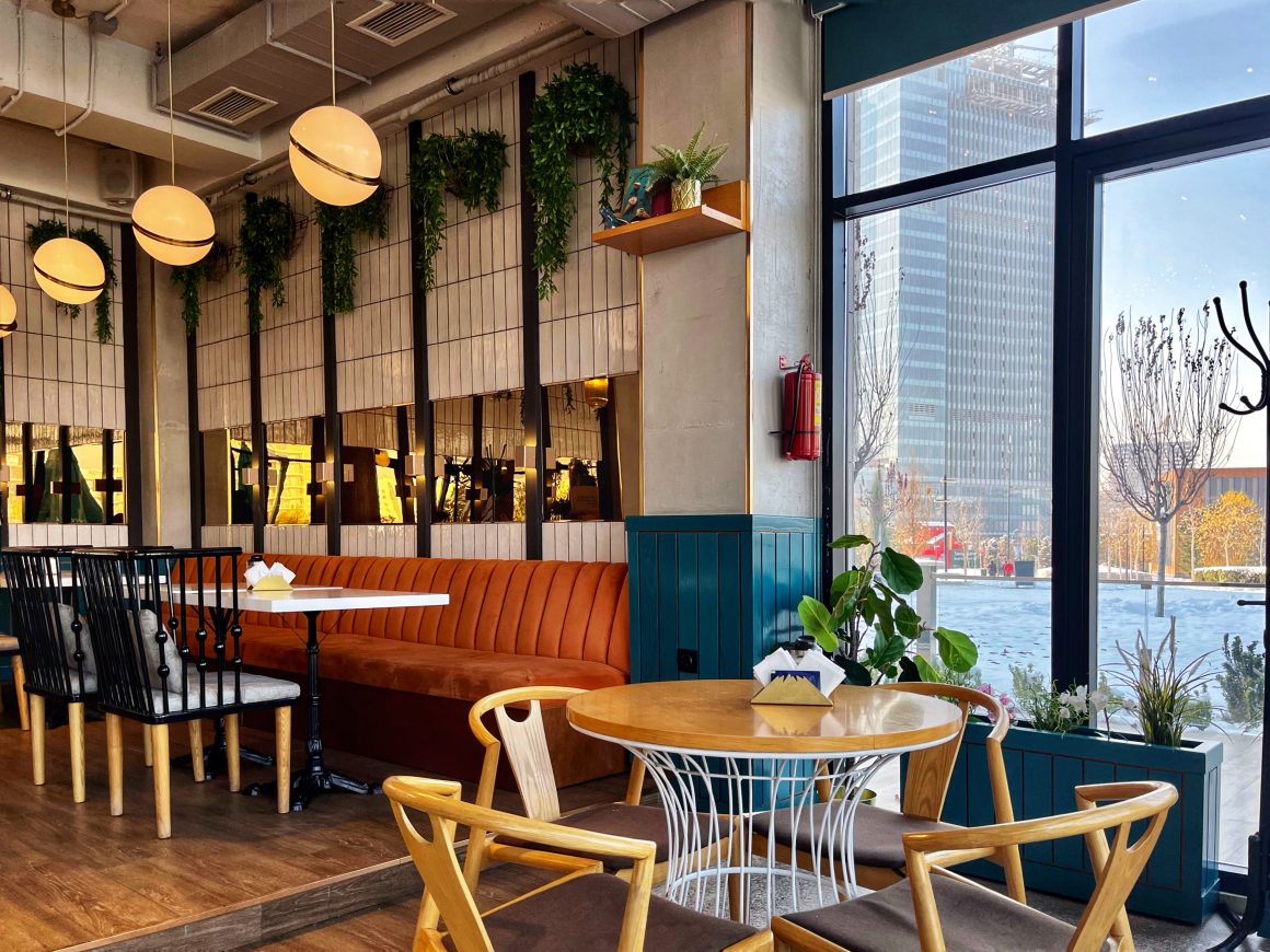 Intérieur restaurant avec banquettes orange, tables en bois, plantes suspendues et grandes baies vitrées
