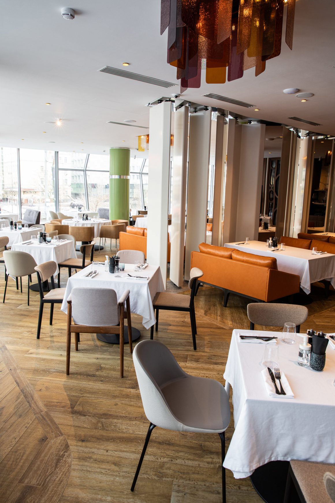 Restaurant moderne avec sol en bois, tables nappées, chaises variées, canapés et grand espace vitré