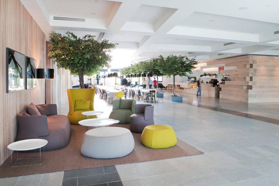 Une grande salle de pause avec canapés gris, chaises jaunes, tables blanches, tapis et plantes