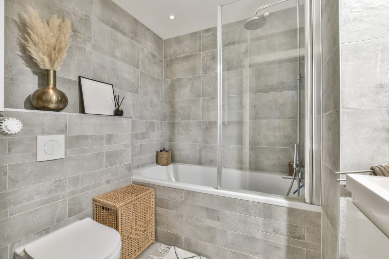 Baignoire vitrée avec carrelage de salle de bain gris texturé, couvrant murs et sol, et accessoires naturels