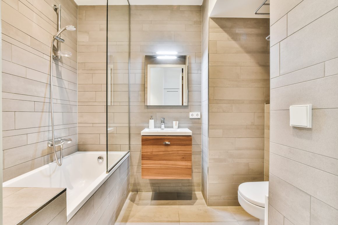 Carrelage de salle de bain beige clair, baignoire avec paroi vitrée, meuble en bois et miroir