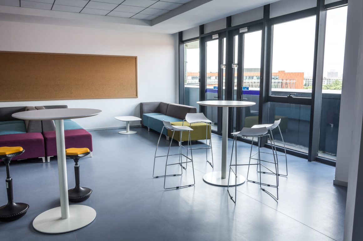 Salle de pause incluant tables rondes, sièges jaunes et blancs, canapé modulable et grand tableau en liège