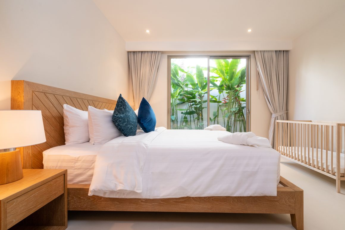 Chambre minimaliste avec lit en bois, draps blancs, coussins bleus, berceau et larges baies vitrées