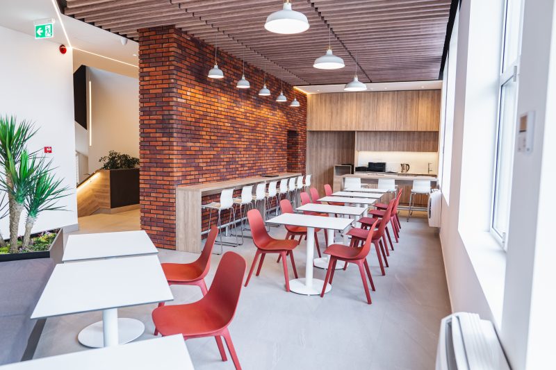 Aménagement réfectoire entreprise avec tables, chaises, comptoir à tabourets, mur en brique et cuisine