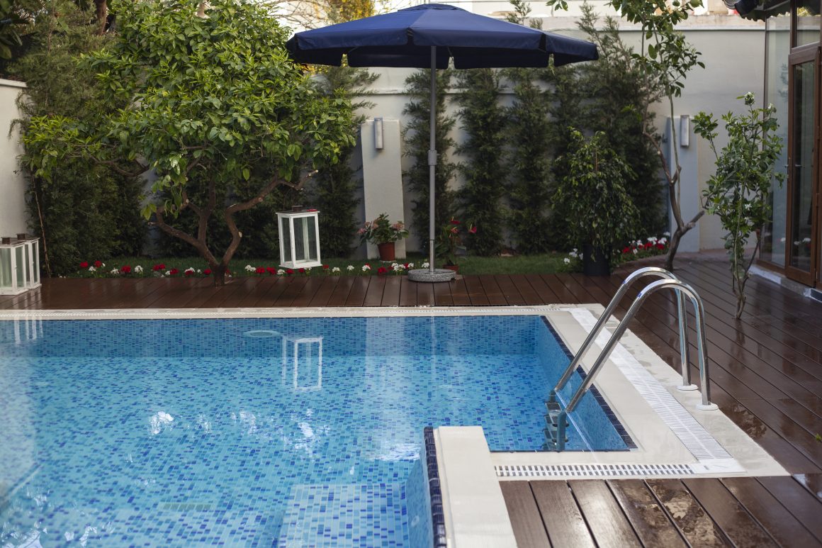 Piscine carrelée en bleu avec margelle blanche dans une terrasse en bois incluant parasol, plantes et lanternes