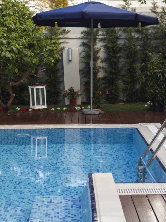 Piscine carrelée en bleu avec margelle blanche dans une terrasse en bois incluant parasol, plantes et lanternes