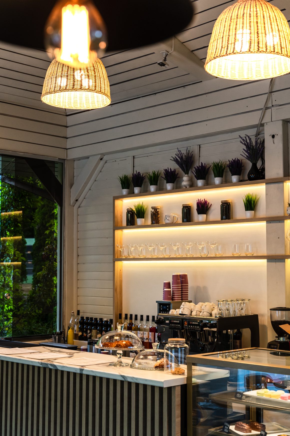 Café chaleureux incluant comptoir élégant, pâtisseries sous cloche et des étagères décorées de plantes