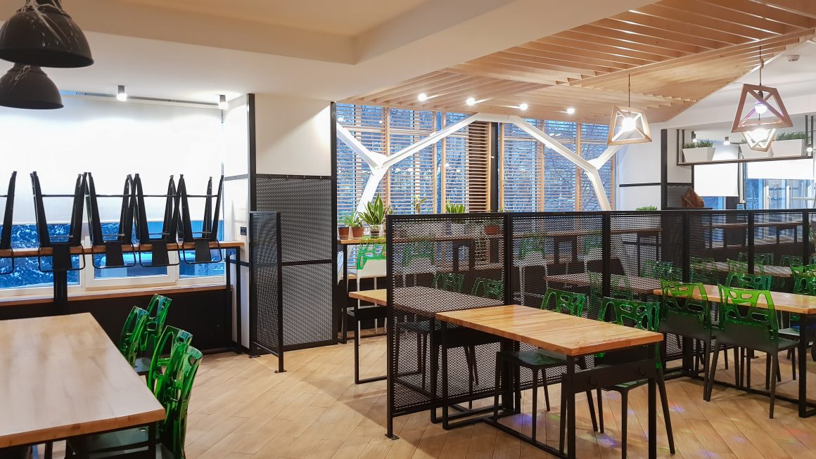 Aménagement réfectoire entreprise avec tables en bois, chaises, cloisons perforées, plantes et plafond lamellé