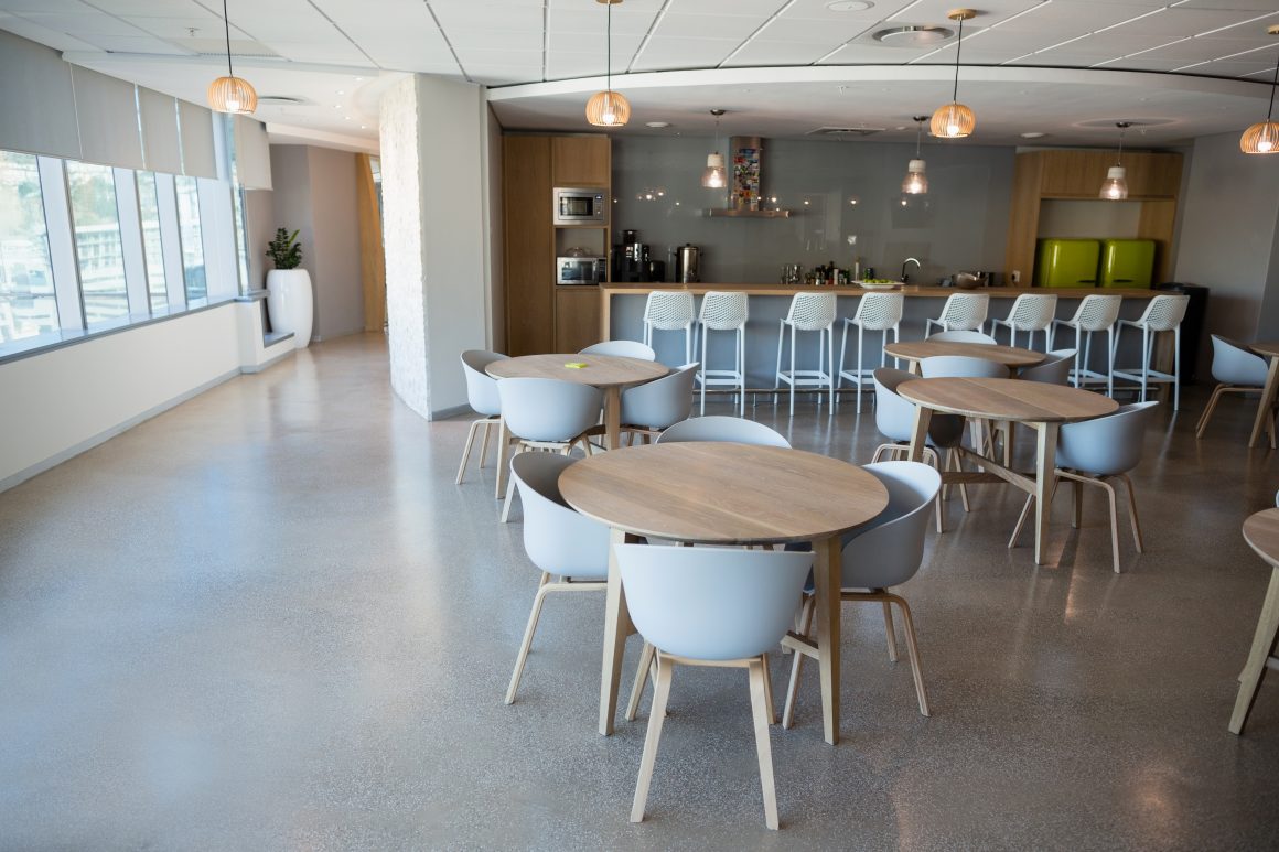 Aménagement réfectoire avec tables rondes en bois, chaises grises, bar avec tabourets et suspensions