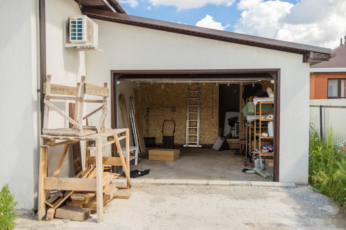 Garage ouvert avec sol en béton, incluant échelles, étagères, palettes en bois, cartons, outils...