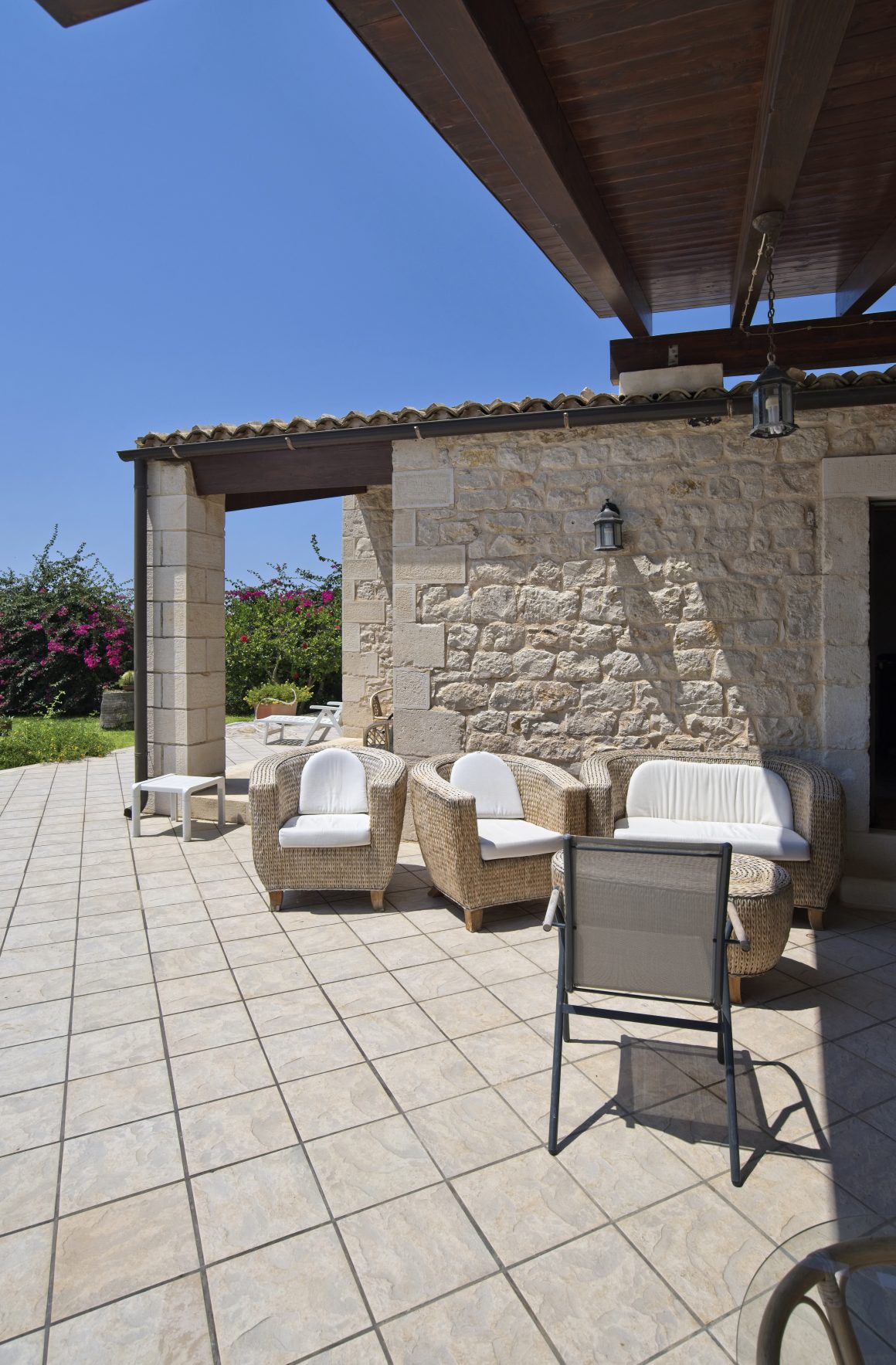 Terrasse ensoleillée avec murs en pierre, fauteuils en rotin blancs, carrelage clair et pergola en bois