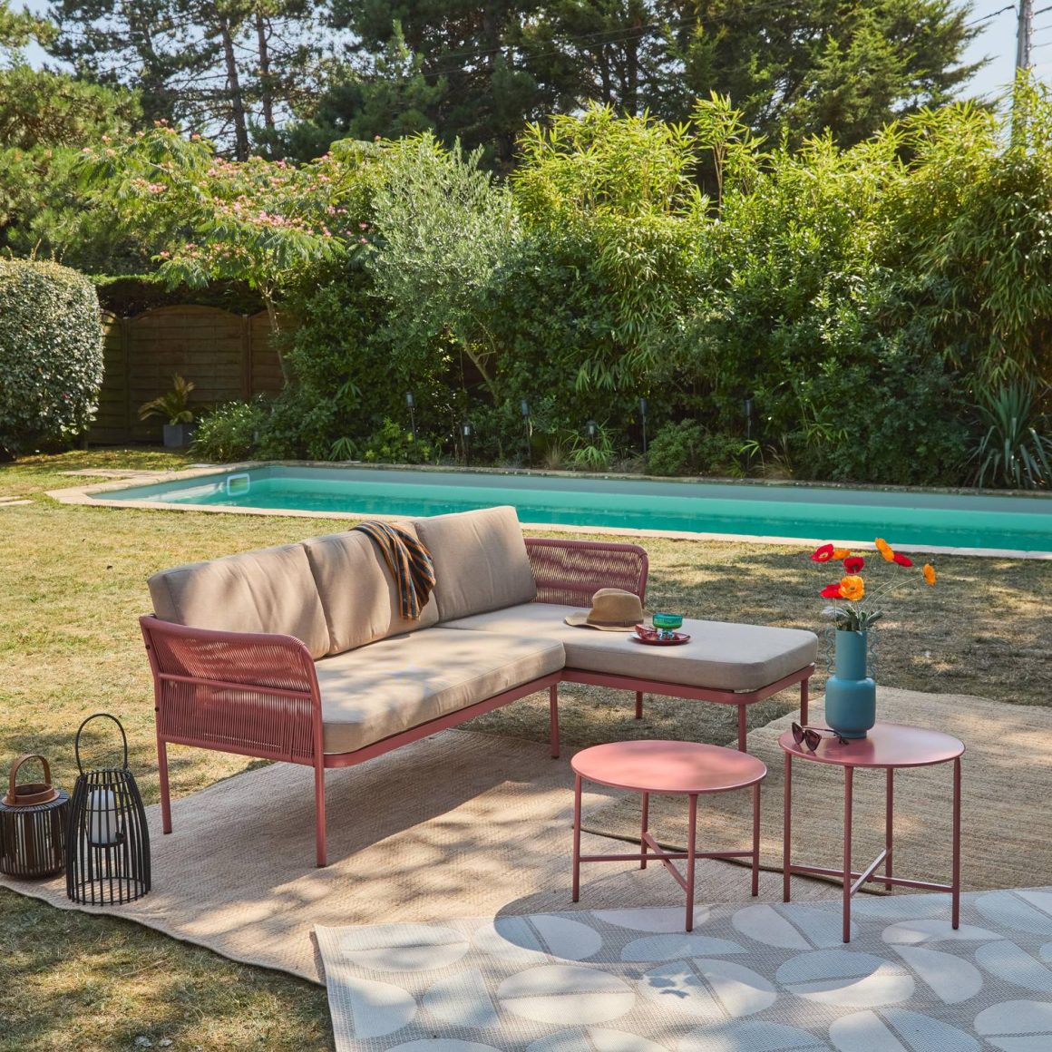 Canapé d'extérieur rose avec coussins beiges, tables basses, tapis, piscine et végétation luxuriante