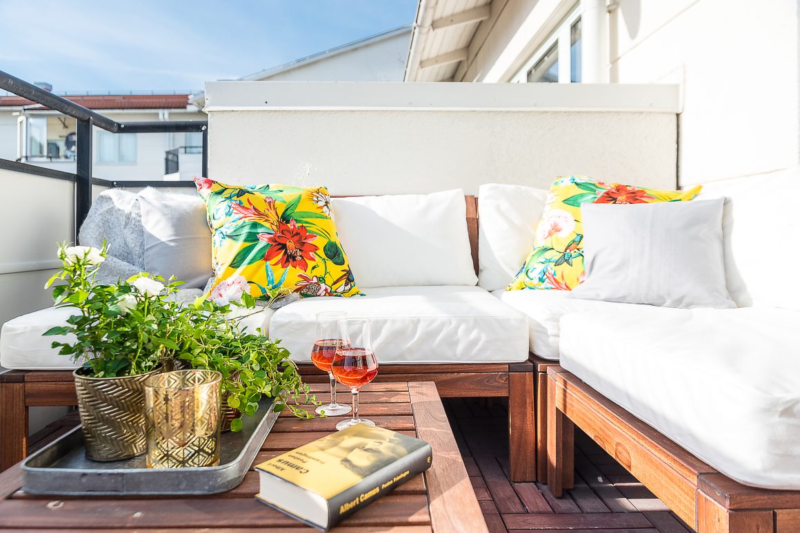 Coin cosy sur balcon : canapé blanc, coussins floraux, table en bois incluant plantes, verres et livre