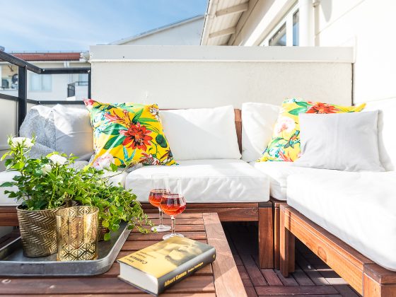 Coin cosy sur balcon : canapé blanc, coussins floraux, table en bois incluant plantes, verres et livre