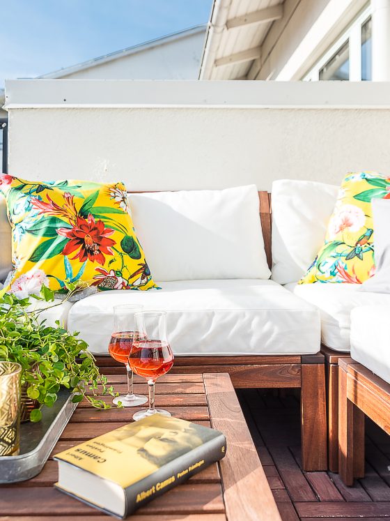Coin cosy sur balcon : canapé blanc, coussins floraux, table en bois incluant plantes, verres et livre