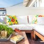 Coin cosy sur balcon : canapé blanc, coussins floraux, table en bois incluant plantes, verres et livre