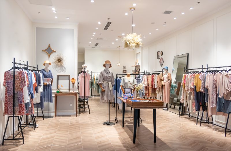 Boutique aménagée avec sol chevrons bois, mannequins, vêtements colorés, miroir, déco murale et luminaires