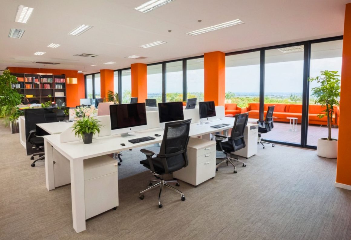 Bureau professionnel avec murs orange, grandes baies vitrées, postes de travail blancs, plantes et coin extérieur avec canapés