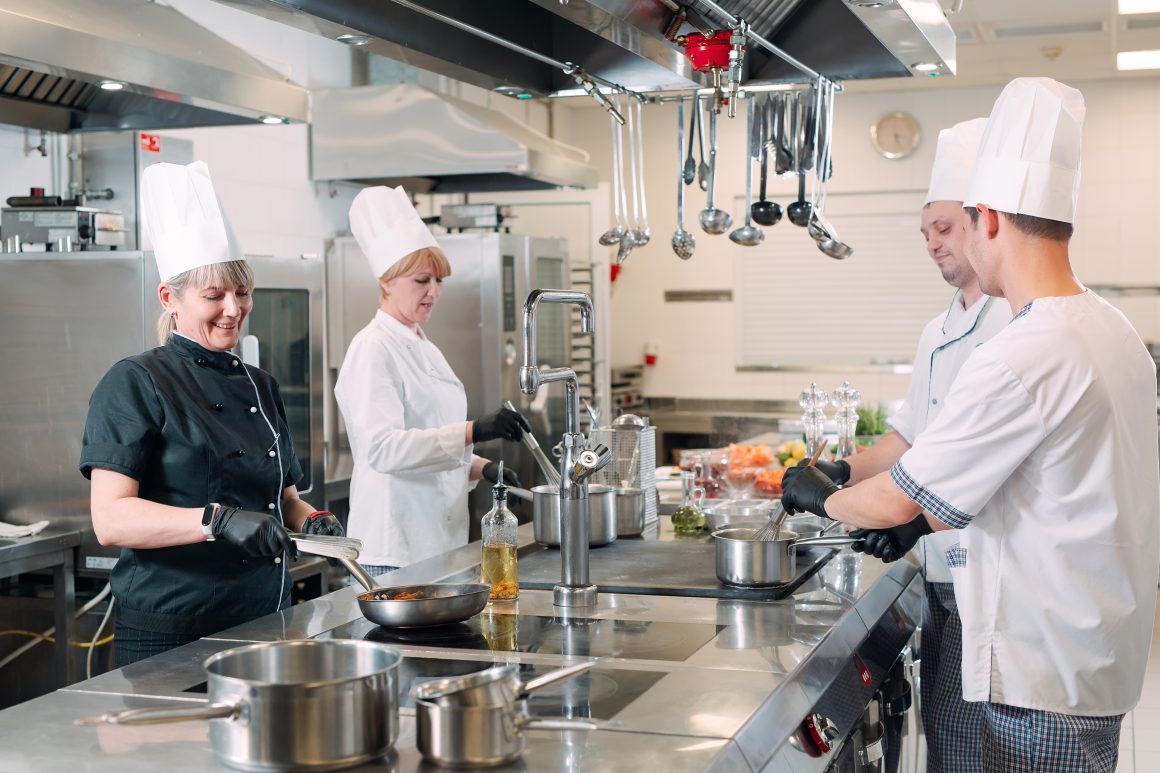 Quatre chefs en cuisine professionnelle, en train de cuisiner autour d'un îlot des ustensiles et robinet