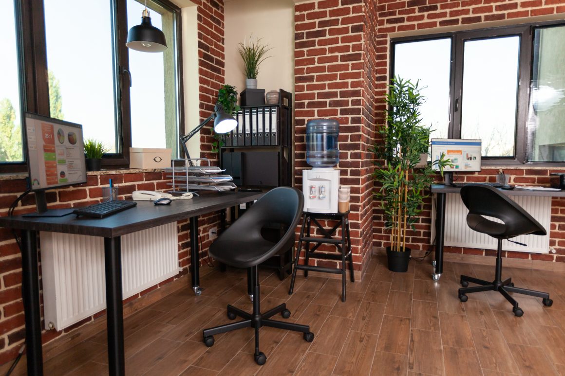 Petit bureau professionnel en briques apparentes aménagé avec deux postes de travail, chaises sur roulettes et sol en bois