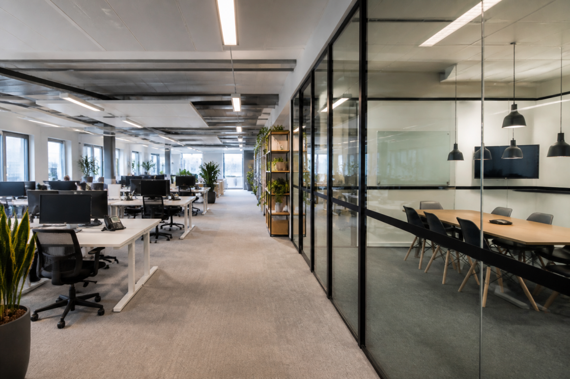 Grand open-space avec bureaux épurés, chaises ergonomiques, plantes et salle de réunion vitrée