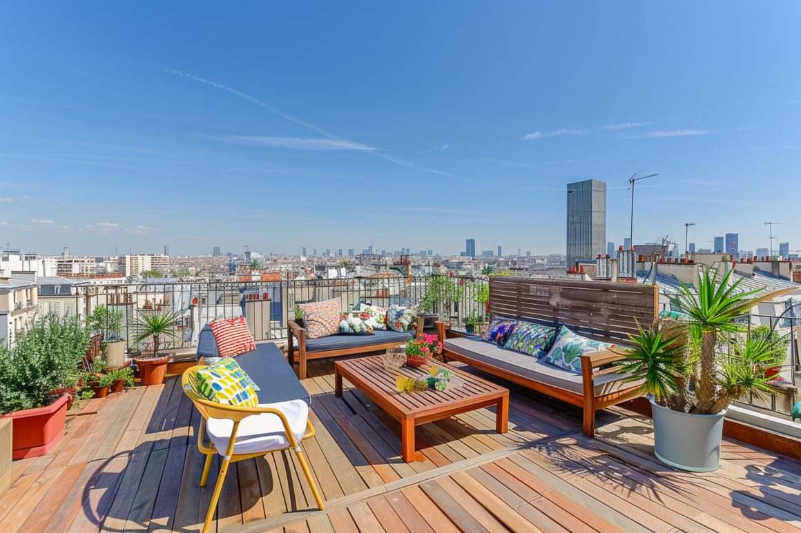 Terrasse sur toit : mobilier en bois, coussins colorés, plantes en pots et vue urbaine lointaine
