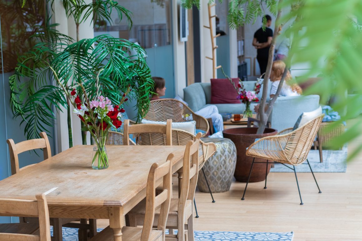 Terrasse café : table en bois, chaises, plantes, fauteuils tressés et personnes discutant dans un coin salon