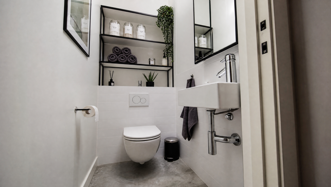 Petit WC suspendu avec carrelage blanc, étagères noires avec serviettes, petit lavabo, miroir et plante