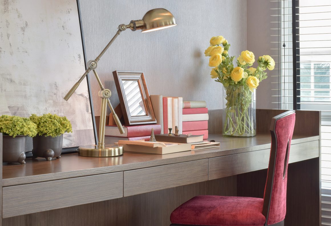Bureau en bois avec lampe dorée, vase de fleurs, chaise velours rouge et fenêtre à stores