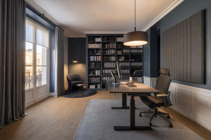 Bureau avec mobilier gris et noir, bibliothèque remplie, panneau acoustique mural et grande fenêtre avec rideaux